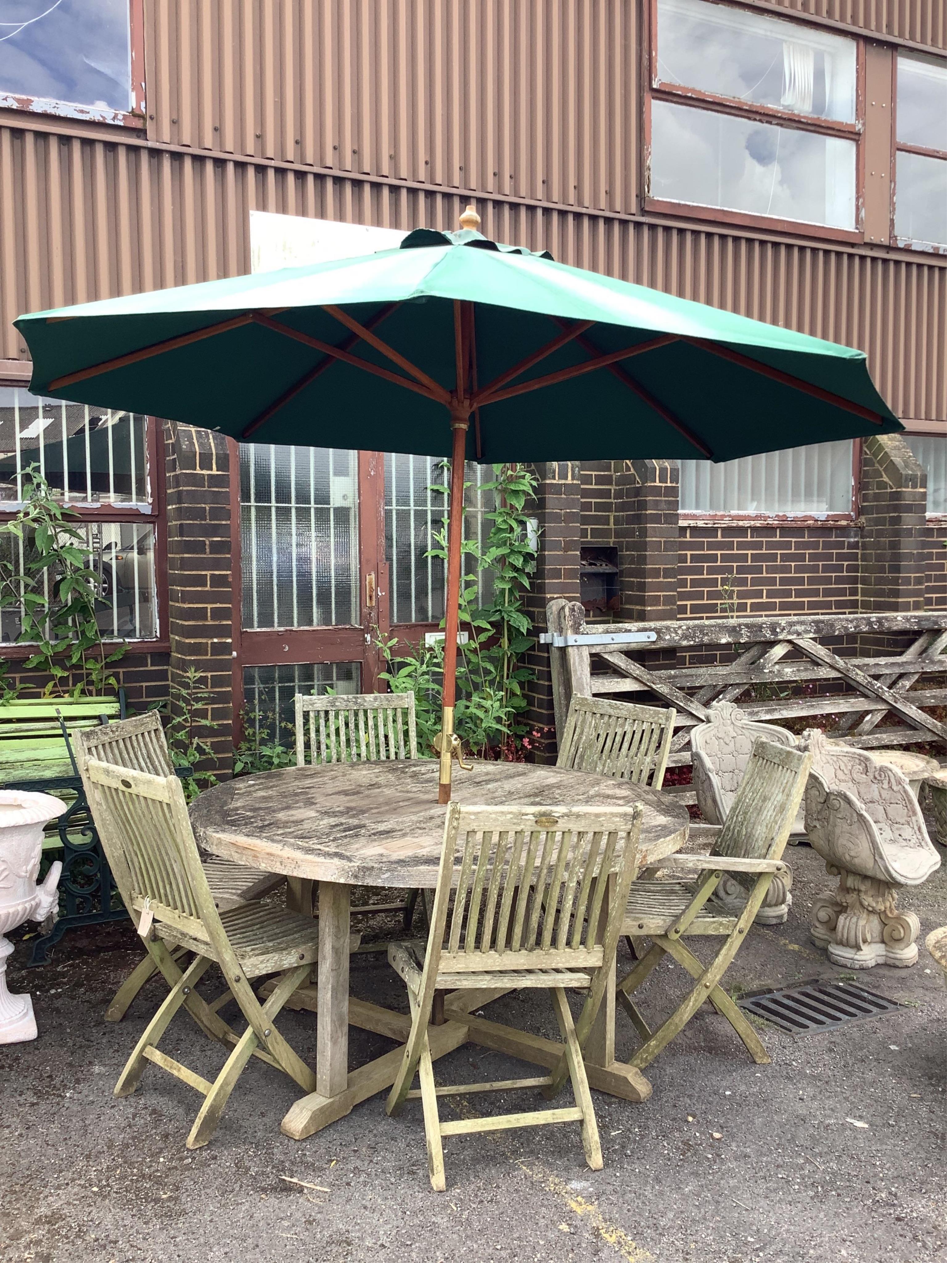 A circular weathered teak garden table, diameter 150cm, height 74cm, together with six teak folding garden chairs, two with arms and a green canvas crank parasol. Condition - fair